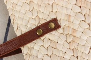 Close-up of handwoven straw bag with dark brown leather strap and vintage metal rivets