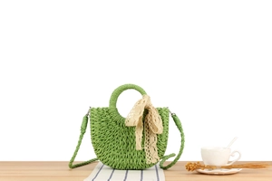 Close-up of green woven semicircular handbag with beige lace bow and wooden surface
