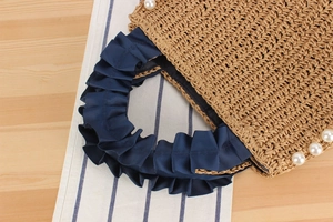 Detail of straw woven bag featuring elegant pearl trim and navy blue pleated ribbon handles