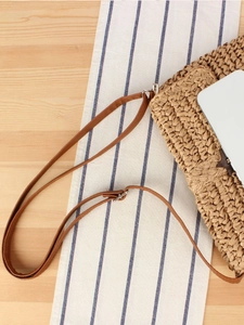 Handwoven straw shoulder bag with adjustable brown leather strap on wooden surface
