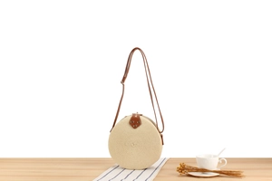 Detail of round straw crossbody bag with brown leather strap on a rustic wooden table