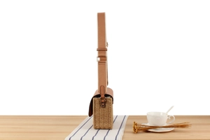 Detail of woven straw bag with brown leather flap and striped strap on a wooden table
