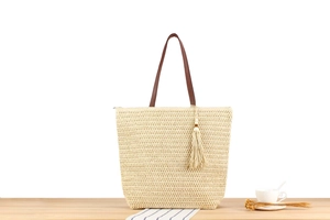 Straw woven tote bag detail showing brown leather straps and decorative tassel on a table