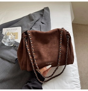 Detail of a brown suede tote bag with chain strap on a white bed alongside accessories