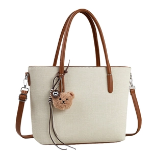 Beige tote bag with brown leather handles and a plush bear charm on a white background