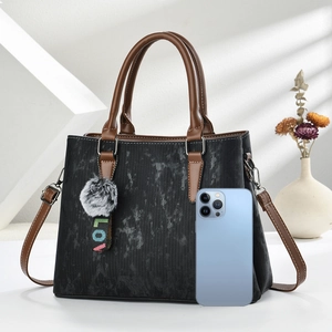 Close-up of black textured handbag with pompom charm beside a smartphone for size reference
