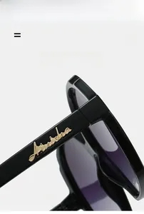 Detailed close-up of golden script logo on black oval sunglasses with purple tinted lenses