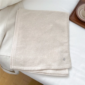 Detailaufnahme eines cremefarbenen Wollschals mit Metalllogo auf einem Bettlaken