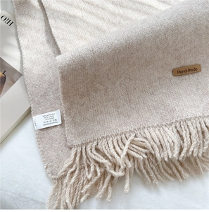 Detail of cream knit scarf showing tassels and handmade leather tag on textured fabric
