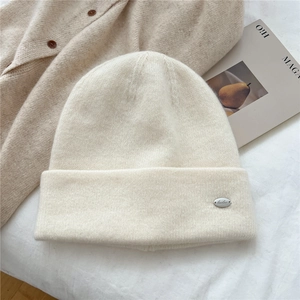 Detail of a creamy white knit beanie featuring a branded silver metal logo plate on the cuff