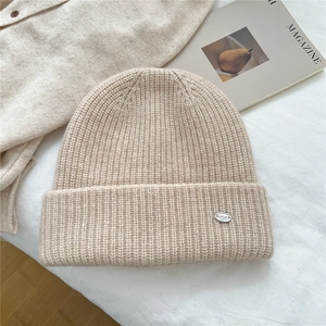 Close-up of beige knit beanie with vertical rib texture and silver oval metal brand tag