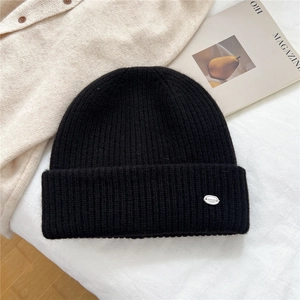 Detail of black ribbed knit beanie with silver metal logo tag on cozy bed setting