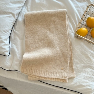 Close-up of beige knit scarf with soft texture and golden metal logo on white bed sheet
