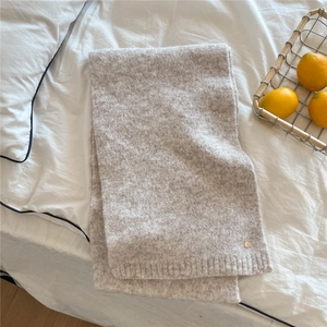 Detail of oatmeal knit scarf with ribbed edge and golden logo on white bedding