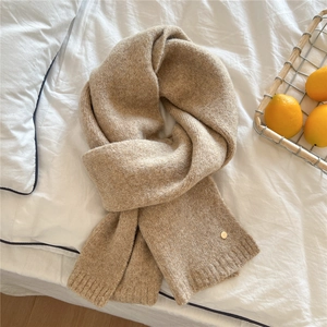 Detail of soft beige knitted scarf with fine texture and golden metal emblem on white linen
