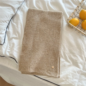 Detail of camel knit scarf with soft ribbed texture and minimalist metal tag on white linen