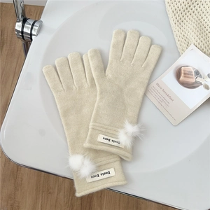 Close-up of beige knit gloves with fluffy white pom-poms and minimalist logo patch