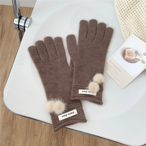 Close-up of brown knit gloves with white pompoms and label on a minimalist setup