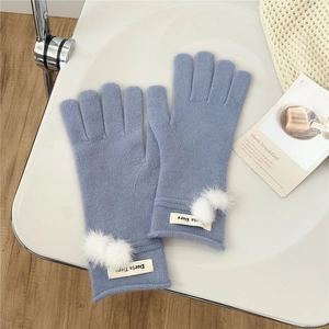 Detail of blue knit gloves with white faux fur pom-poms and a minimalist logo tag