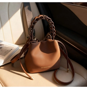Detail of a brown leather bucket bag featuring a thick dark brown woven handle on a car seat