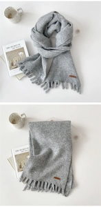 Detail view of a light grey knit scarf featuring fringe edges and a brown leather logo tag