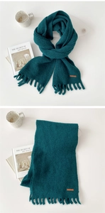 Close-up of a dark green knit scarf featuring short tassels and a brown leather logo patch