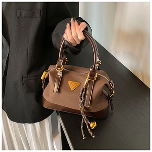 Detail einer braunen Boston Bag mit goldener Dreiecksplakette und geflochtenem Anhänger