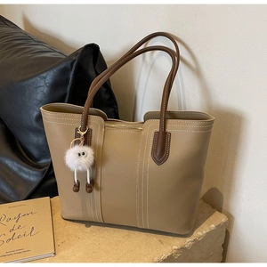 Detail of khaki tote bag with contrast stitching and plush monster charm on stone surface