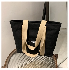 Detail of a black canvas tote bag with beige straps and white letter logo on a glass table
