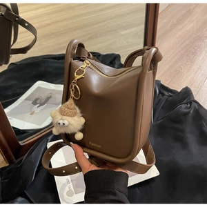 Detail of a brown leather crossbody bag featuring a golden zipper and a cute plush monster charm