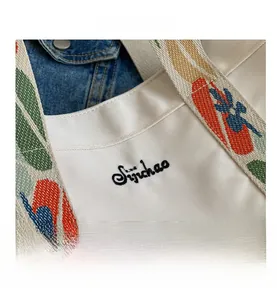 Detail of white canvas tote bag featuring black letter embroidery and colorful striped strap