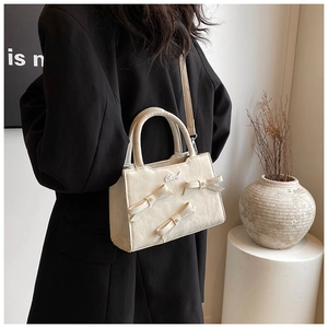 Detail der cremefarbenen Handtasche mit 3D-Schleifen und Logo vor schwarzem Blazer
