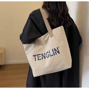 Close-up of a large beige canvas tote bag with blue logo text on a model's shoulder