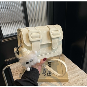 Detail of a beige crossbody bag featuring large decorative buckles and a cute plush charm