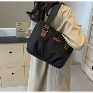 Detail of a large black canvas tote bag with brown leather trim worn by a model in a minimalist room