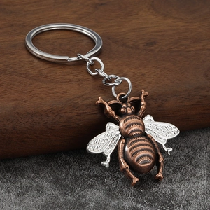 Minimalist bronze bee keychain with silver wings on a rustic wooden and stone background