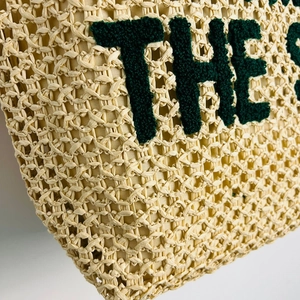 Detailed close-up of woven straw texture and dark green embroidered lettering on tote bag