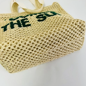 Detail of beige straw tote bag with green embroidered The Sun letters and textured weave