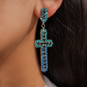 Gros plan d'une boucle d'oreille croix en strass bleu et vert portée par un mannequin en détail.