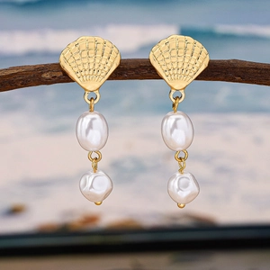 Elegant gold shell earrings with baroque pearls hanging on wood with a beach backdrop