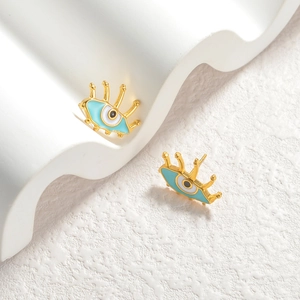 Golden evil eye stud earrings with blue enamel and eyelash details on white display