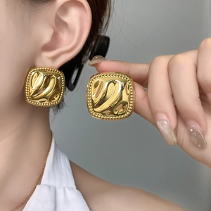 Detail of square gold earrings featuring wavy texture and beaded edge on model ear