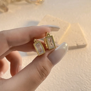 Elegant gold-plated square zircon earrings held by hand against a soft warm stone backdrop