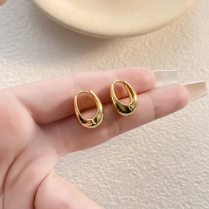 Minimalist gold-plated oval hoop earrings held by hand for scale on warm background