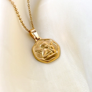 Collier en acier doré avec pendentif pièce ange en relief posé sur du tissu en soie