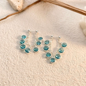 Detail of silver drop earrings with mint blue zircon tassels on a textured beige surface
