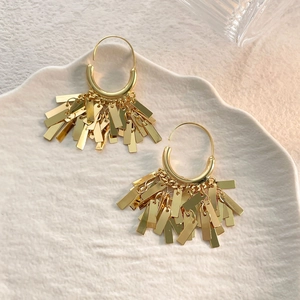 Close-up of gold fringe earrings with hanging metallic plates on a white ceramic plate