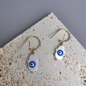 Gold Hamsa hand shell earrings with blue evil eye detail on stone background