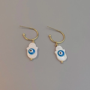 Macro view of gold Hamsa shell earrings with blue evil eye detail on grey background