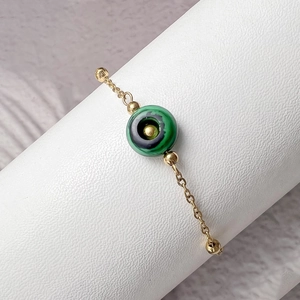 Macro shot of gold-plated evil eye bracelet with green stone detail on a white pedestal